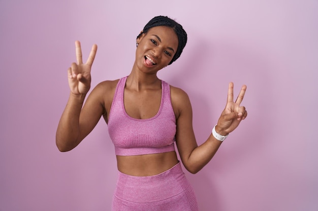 Free photo african american woman with braids wearing sportswear over pink background smiling with tongue out showing fingers of both hands doing victory sign. number two.
