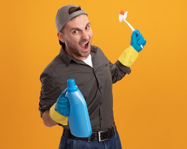 Free photo angry young cleaning man wearing casual clothes and cap in rubber gloves holding cleaning brush and bottle with cleaning supplies shouting with aggressive expression standing over orange wall