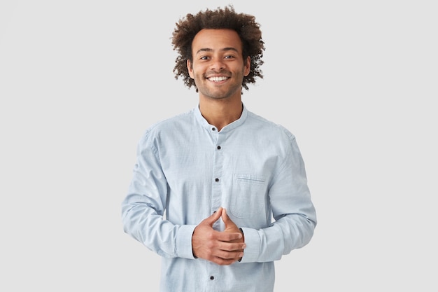 Free photo attractive mixed race male with positive smile, shows white teeth, keeps hands on stomach, being in high spirit, wears white shirt, rejoices positive moments in life. people and emotions concept