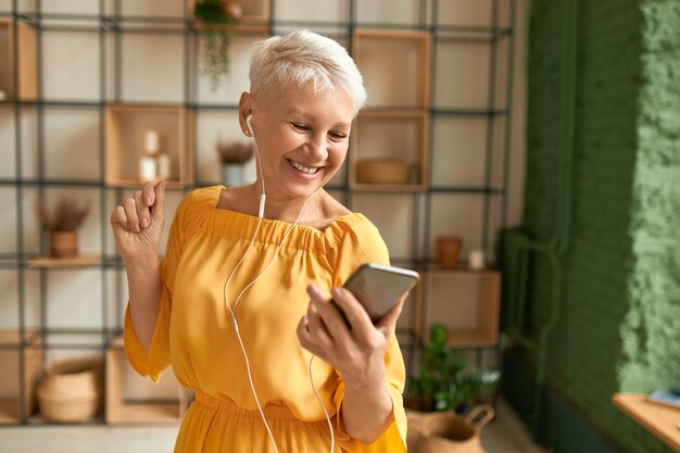 Attractive cheerful female pensioner in yellow dress using mobile phone, listening to music in earphones, dancing, having happy joyful facial expression