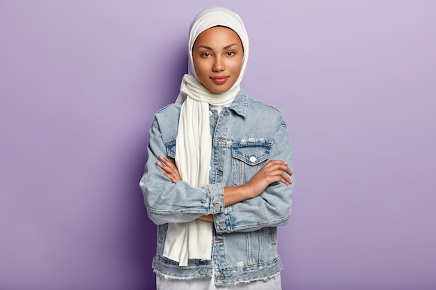 Free photo attractive eastern woman covers head with white headscarf to guard her dignity and power, has special dress code, keeps hands crossed, looks with modesty, poses over purple wall. islamic rules