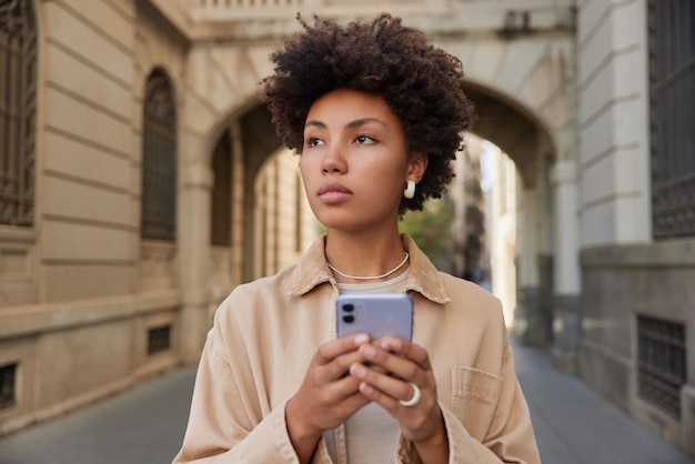 Бесплатное фото Привлекательная модель читает текстовое сообщение на смартфоне, прогуливается во время пешеходной экскурсии на свежем воздухе, имеет задумчивое выражение лица, наслаждается отдыхом, позирует возле древнего здания молодой турист с телефоном