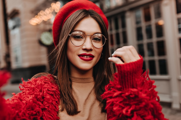 Бесплатное фото Привлекательная женщина с красными губами делает селфи на улице. выстрел из брюнетки в очках, одетой в стильную шляпу, красный пиджак и бежевый топ.