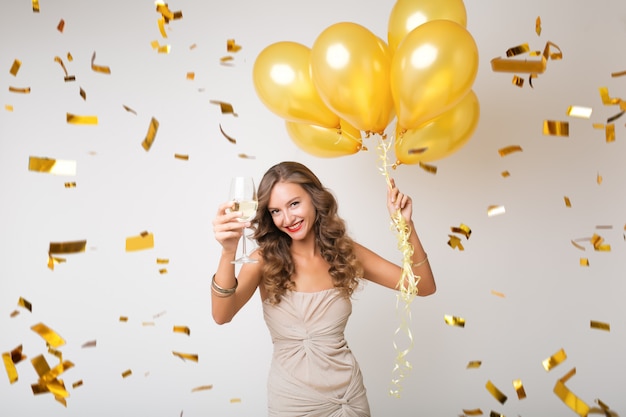 Бесплатное фото Привлекательная молодая стильная женщина празднует новый год, пьет шампанское, держит воздушные шары, летит золотое конфетти, улыбается счастливым, изолированным, в праздничном платье