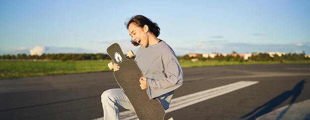 무료 사진 아름다운 아시아 십대 소녀가 기타를 연주하는 것처럼 스케이트보드를 들고 롱보드를 가지고 놀고 있습니다.