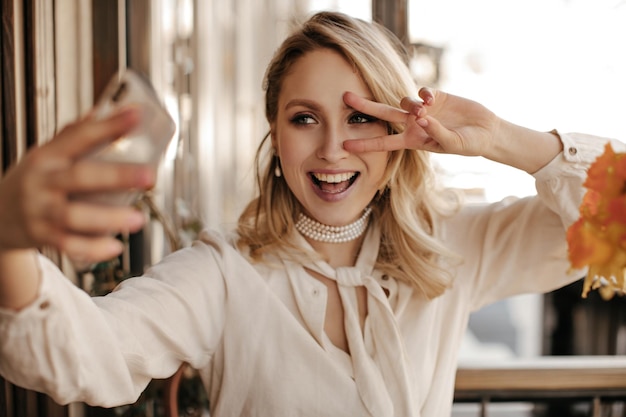 Бесплатное фото Красивая блондинка в жемчужном ожерелье и стильной белой блузке показывает знак мира, улыбается и делает селфи в ресторане