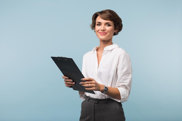 Бесплатное фото Красивая деловая женщина с темными короткими волосами в белой рубашке держит в руках черную папку, радостно глядя в камеру на синем фоне