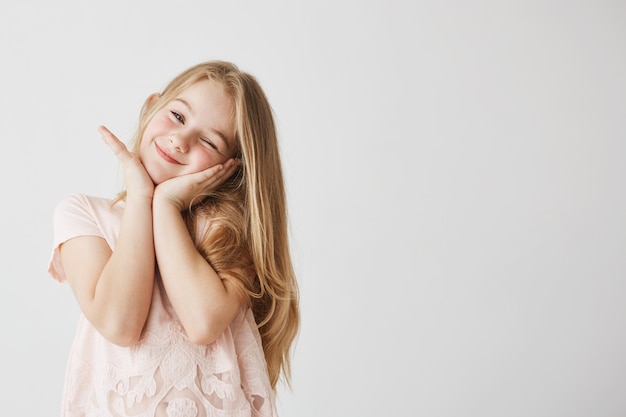 Бесплатное фото Красивая маленькая блондинка улыбается, подмигивая, позирует, трогательно лицо руками в розовом милое платье. ребенок смотрит счастливым и восхищенным копировать пространство