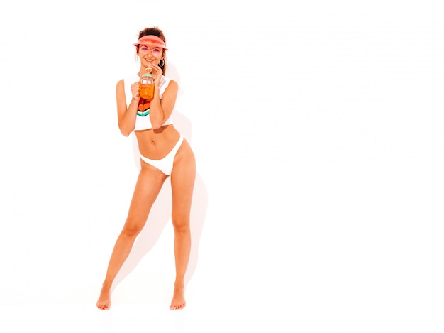 Beautiful smiling sexy woman in white summer underpants and topic. Trendy girl in sunglasses and transparent visor cap. Going crazy. Funny model isolated. Drinking fresh cocktail smoozy drink