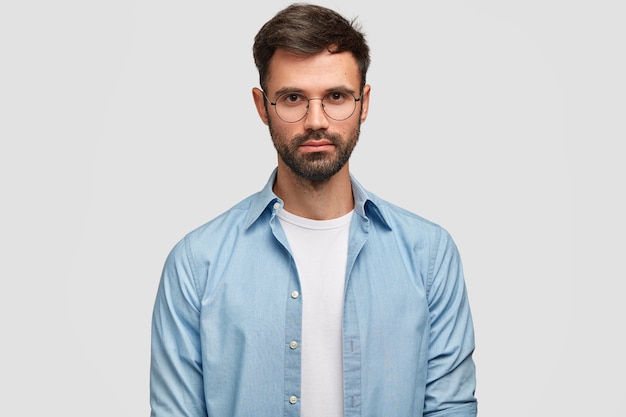 Free photo bearded young self confident male with pleasant appearance, dressed in blue shirt, looks directly, isolated over white wall. handsome man freelancer thinks about work indoor.