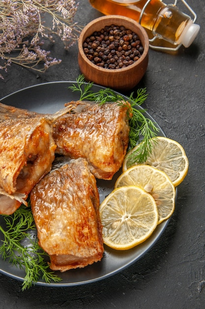 Вид снизу жареная рыба с ломтиками лимона на тарелке черный перец в деревянной миске бутылка масла на темном фоне