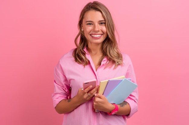Бесплатное фото Блондинка в розовой рубашке улыбается, держа блокноты и используя смартфон