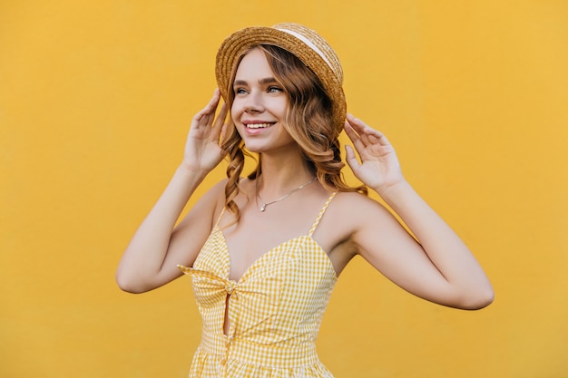 Бесплатное фото Беспечная белая девушка в соломенной шляпе, мечтательно глядя в сторону. портрет утонченной красивой дамы в стильном желтом платье.