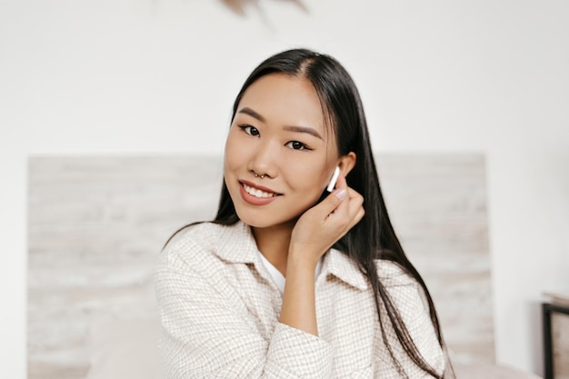 Free photo browneyed brunette asian woman in good mood listens to music in wireless headphones and smiles sincerely portrait of attractive lady in beige jacket looks into camera in white room