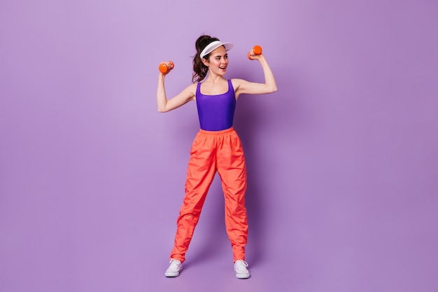 Бесплатное фото Брюнетка в кепке занимается фитнесом на фиолетовой стене