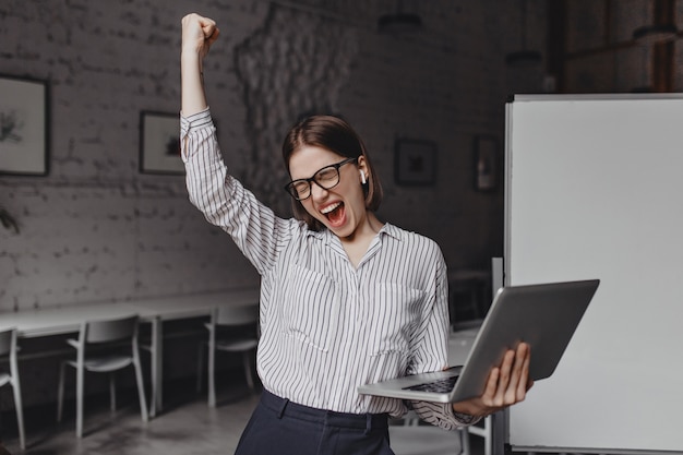 Free photo business woman with laptop in hand is happy with success. portrait of woman in glasses and striped blouse enthusiastically screaming and making winning gesture.