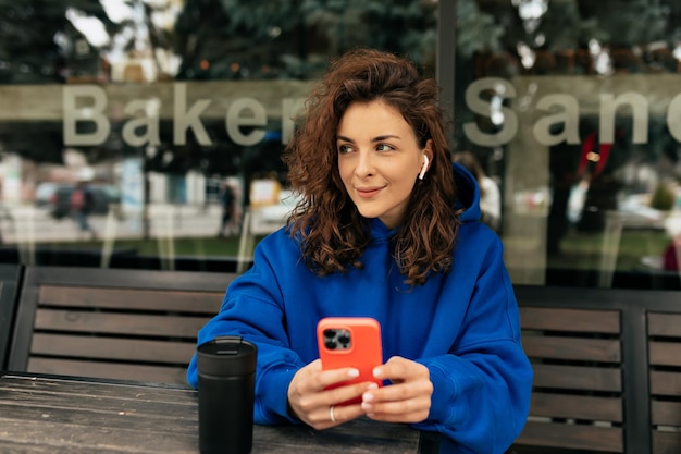 Бесплатное фото Беззаботная молодая европейская девушка использует современный смартфон, сидя на улице с кофе кудрявая брюнетка носит пуловер и джинсы концепция технологии