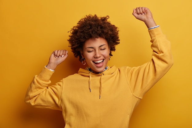 Free photo carefree energetic dark skinned young woman dances with hands up, sings favorite song, triumphs over win, closes eyes, expresses happiness, achieves victory or approval, wears yellow sweatshirt
