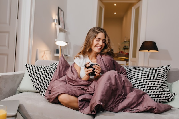 Бесплатное фото Жизнерадостная женщина сидит на диване с одеялом и подушками и улыбается