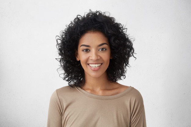 Free photo cheerful dark-skinned woman smiling broadly, rejoicing at her victory in competition among young writers, standing isolated against grey wall. people, success, youth and happiness concept