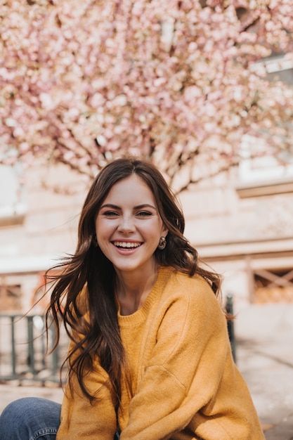 Foto gratuita ragazza allegra in maglione di cachemire ride sullo sfondo di sakura sbocciante. ritratto di donna in felpa con cappuccio gialla in città in primavera