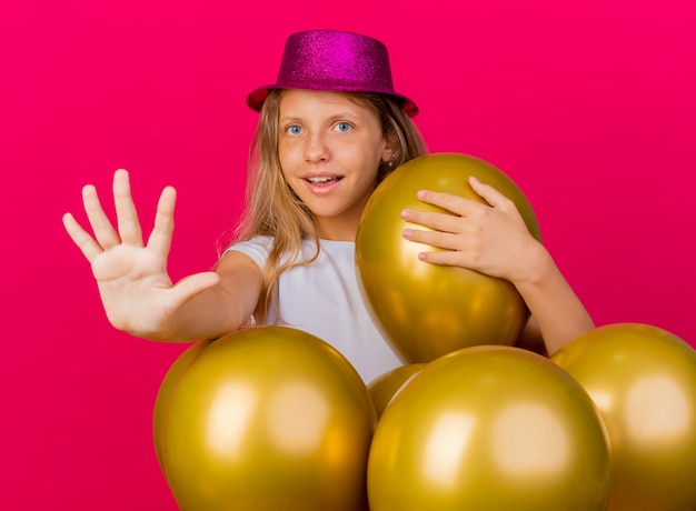 Бесплатное фото Веселая симпатичная маленькая девочка в праздничной шляпе с кучей воздушных шаров, глядя в камеру, делая знак сто с улыбкой руки, концепция вечеринки по случаю дня рождения, стоящая на розовом фоне