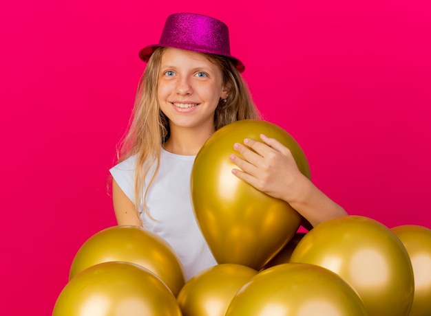 Бесплатное фото Веселая симпатичная маленькая девочка в праздничной шляпе с кучей воздушных шаров, глядя в камеру, улыбаясь со счастливым лицом, концепция вечеринки по случаю дня рождения, стоящая на розовом фоне