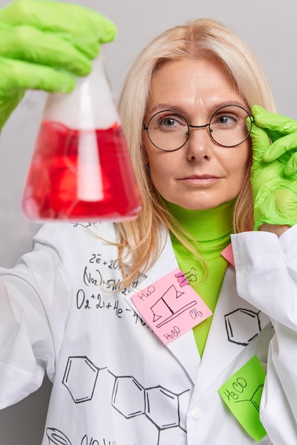 Бесплатное фото Химик проводит химический тест научных исследований в лаборатории, держит фляжку с красной жидкостью, носит очки, белый халат позирует в помещении. биохимия или фармацевтические разработки