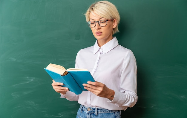 Бесплатное фото Концентрированная молодая блондинка учительница в очках в классе, стоя перед классной доской, держа и читая книгу