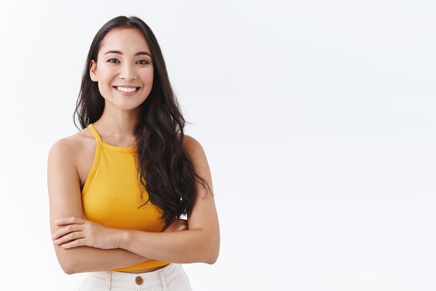 Free photo confident, attractive young outgoing asian woman in yellow top, smiling friendly and happy as cross hands chest, posing over white background self-assured, sassy pose, look determined