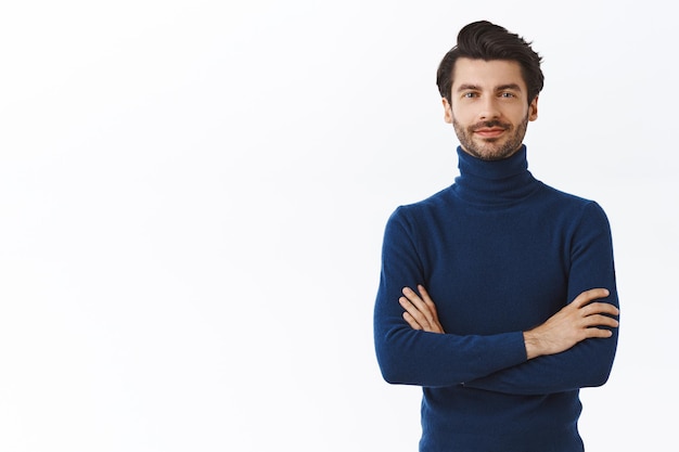 Бесплатное фото Уверенный в себе красивый мужчина-предприниматель в синем свитере с высоким воротом, скрестив руки на груди, дерзкая и самоуверенная ухмылка.