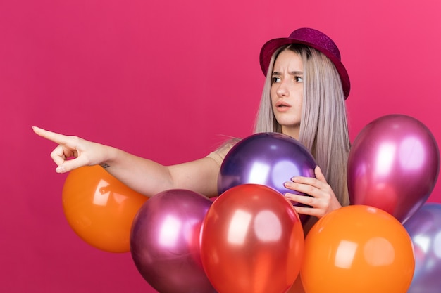 Бесплатное фото Смущенная молодая красивая девушка в партийной шляпе с зубными скобами, стоящая за воздушными шарами, сбоку изолирована на розовой стене