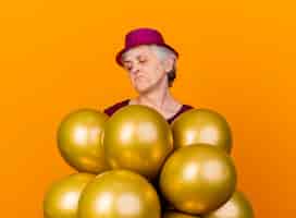 Бесплатное фото Смущенная пожилая женщина в партийной шляпе держит и смотрит на гелиевые шары, изолированные на оранжевой стене с копией пространства