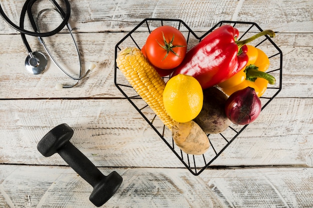 Free photo colorful raw vegetables in container near dumbbell and stethoscope on wooden surface