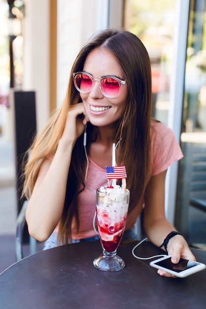 Close-up of cute girl sitting in a cafe eating ice-cream with cherry on top. She wears pink top and pink eyeglasses. She listens to music on smartphone and smiles. She has long dark hair