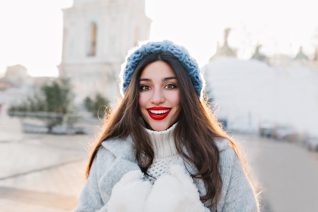 흐림시에 웃 고 붉은 입술으로 갈색 머리 여자의 클로즈업 초상화. 파란색 니트 모자와 놀란 얼굴 표정으로 포즈를 취하는 따뜻한 장갑에 평온한 소녀의 야외 사진.