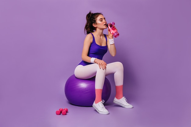 Бесплатное фото Кудрявая темноволосая женщина в спортивной одежде в стиле 80-х сидит на фитболе и пьет воду из розовой бутылки