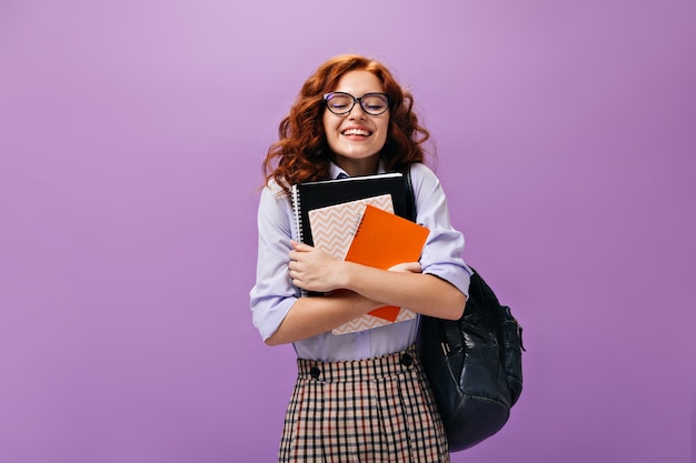 Бесплатное фото Кудрявая девушка в очках держит тетради на фиолетовой стене