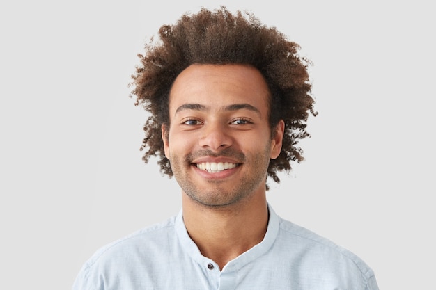 Free photo curly man with broad smile, shows perfect teeth, being amused by interesting talk, has bushy curly dark hair stands indoor against white blank wall