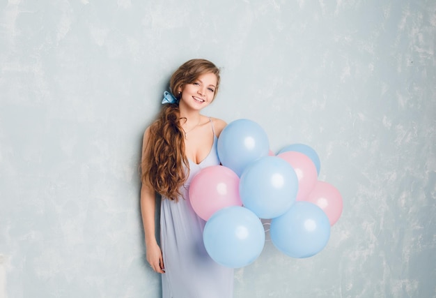 Бесплатное фото Милая белокурая девушка, стоя в студии, улыбаясь и держа синие и розовые воздушные шары. она носит светло-голубое платье и длинные волнистые волосы, заплетенные в косу, с голубой лентой.