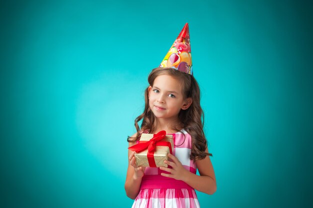 Милая веселая маленькая девочка с подарком и праздничной шапочкой на синем фоне