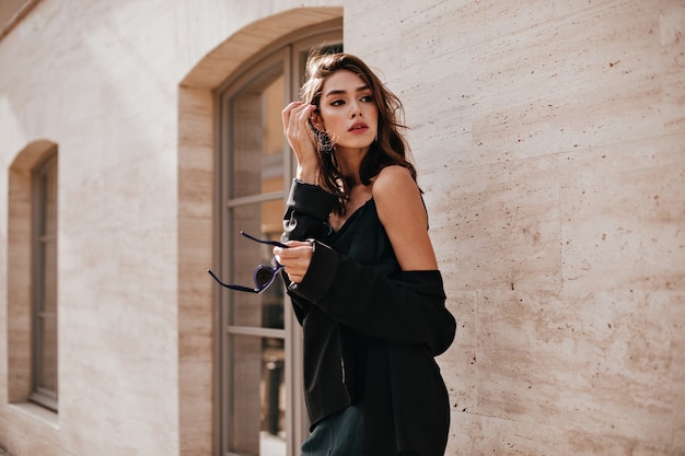 Бесплатное фото Симпатичная молодая девушка с темной волнистой прической и ярким макияжем, шелковое платье, черный пиджак, держит в руках солнцезащитные очки и смотрит в сторону на бежевую стену здания