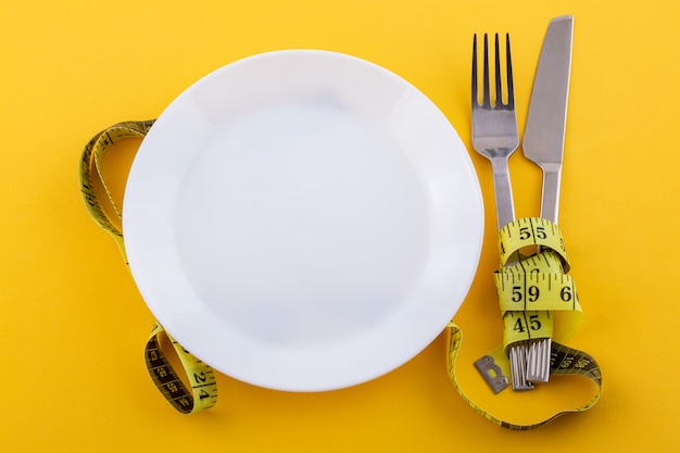 Free photo cutlery and a white plate with measuring tape on a yellow, the concept of weight loss and diet
