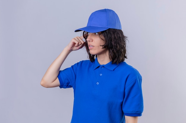 Foto gratuita ragazza delle consegne in uniforme blu e cappuccio che guarda lontano con espressione confusa in piedi sopra lo spazio bianco isolato