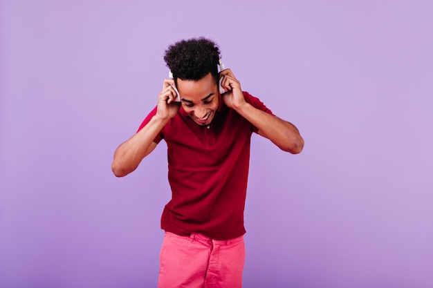 Free photo ecstatic black young man touching his headphones and looking down. indoor photo of jocund male model in red attire.