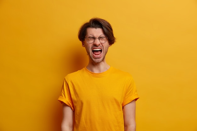 Free photo emotional guy keeps mouth wide opened, screams from disappointment, shouts with closed eyes, feels troubled as lost huge bet, dressed in bright yellow t shirt, looses temper, has life problems