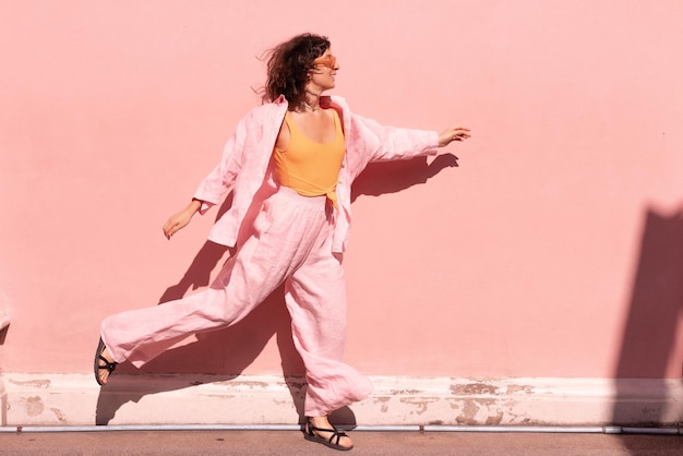 Free photo energetic young caucasian woman in loose clothes moves with flying gait along pink wall on sunny day