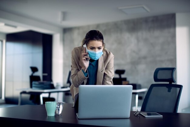 Entrepreneur with face mask using laptop and communicating over mobile phone at her office