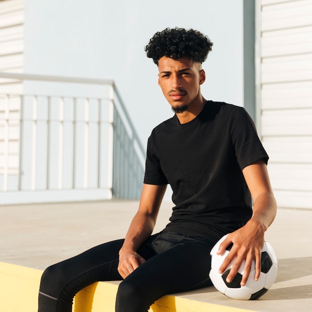 Этнический спортивный парень сидит с футбольным мячом