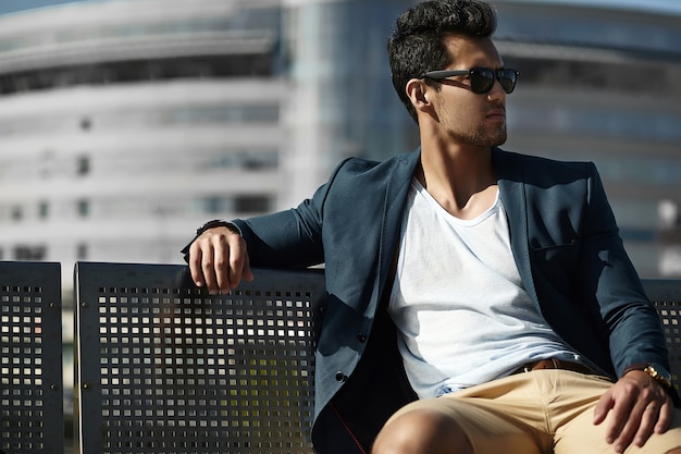 Free photo fashion portrait of young sexy businessman handsome model man in casual cloth suit in sunglasses sitting on a bench in the street